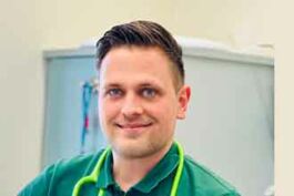 Person mit kurzem Haar, grünem T-Shirt, Stethoskop um den Hals, lächelnd.