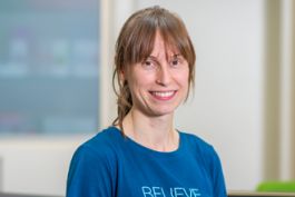 Person, hellbraunes Haar, lächelnd, blaues T-Shirt mit weißem Textaufdruck, unscharfer Hintergrund.