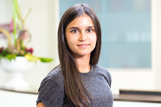 Frau mit langen, dunklen Haaren; graues T-Shirt; neutraler Gesichtsausdruck; verschwommener, heller Hintergrund.