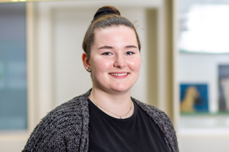 Person, braunes Haar im Dutt, grauer Cardigan, schwarzes Oberteil, lächelt, neutraler Hintergrund.